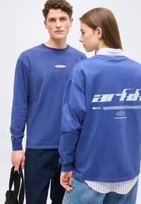 Νεαρός άνδρας και γυναίκα που φορούν μπλε πουκάμισα με λευκό κείμενο; ο άνδρας κρατάει μία μαύρη τσάντα, η γυναίκα φοράει ριγέ πουκάμισο από μέσα, στέκονται δίπλα-δίπλα.