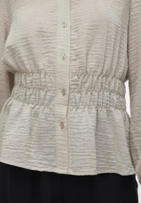Lys beige bluse i teksturert stoff, med en tettsittende midje med elastikk, knappelukking foran, og milde volanger i kanten.