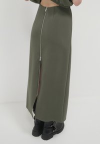 Gonna maxi verde oliva in un tessuto liscio, con chiusura a zip sul retro e uno spacco alto. Abbinata a stivali neri alla caviglia.