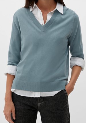 Woman wearing light blue V-neck sweater over white button-up shirt with rolled sleeves and black jeans, standing with one hand in pocket.