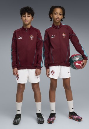 Kaksi nuorta urheilijaa pukeutuneina viininpunaisiin Portugalin jalkapallotakkeihin, valkoisiin shortseihin ja nappulakenkiin; toinen heistä pitelee Portugal-teemalla varustettua jalkapalloa.