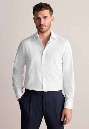 Mand med kort brunt hår iført en hvid skjorte med knapper og mørkeblå bukser, står med den ene hånd i lommen.