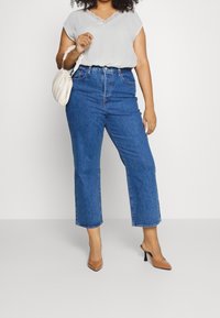Kvinde iført hvid bluse, blå højtaljede cropped jeans, brune spidse hæle og holder en lille hvid håndtaske på ensfarvet baggrund.