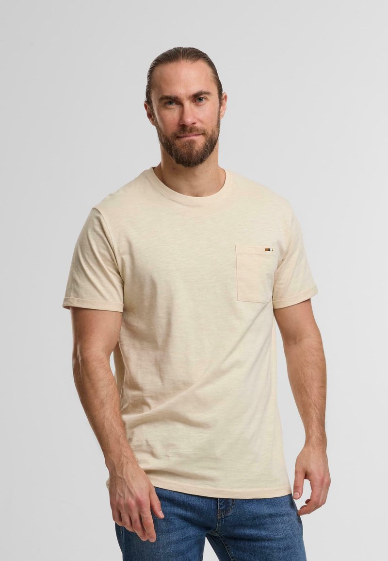 Beige cotton t-shirt with a round neck, short sleeves, and a left chest pocket. Slightly textured fabric, paired with blue jeans.