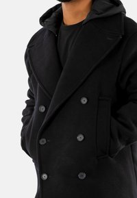 Abrigo negro de corte ajustado y double-breasted, con grandes botones, solapas en pico y una capucha desmontable fabricada en material textil.