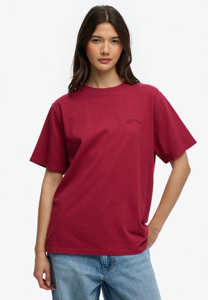 Młoda kobieta z długimi ciemnymi włosami, ubrana w prosty czerwony T-shirt z krótkim rękawem i jasnoniebieskie dżinsy, stoi z jedną ręką w kieszeni.