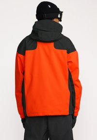 Veste orange et noire avec capuche, présentant un tissu texturé, des poignets élastiques et un design élégant, principalement visible de dos.