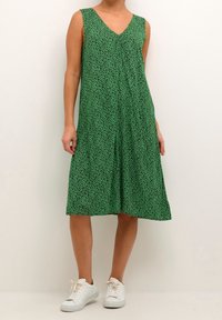 Vestido verde sin mangas con escote en V, de tejido texturizado con un patrón de leopardo negro, que llega hasta la media pantorrilla, combinado con zapatillas blancas.