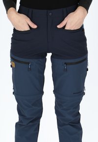 Navy blue pants with a multi-panel design, featuring front pockets and zippered thigh pockets with contrasting zippers and branding detail.