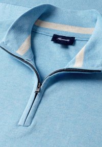 Jasnoniebieski sweter z dzianiny z białymi paskami wewnątrz kołnierza oraz częściowo rozpinanym srebrnym zamkiem z przodu przy dekolcie.