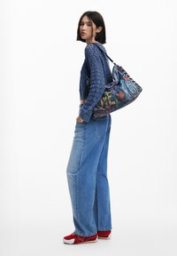 Junge Frau in blauer Strickjacke und weiter Jeans, die eine bunte Schultertasche im Graffiti-Stil trägt, mit roten Turnschuhen, seitlich stehend.