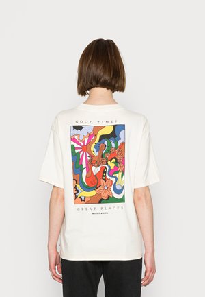 Person mit kurzen braunen Haaren, die ein übergroßes weißes T-Shirt mit buntem abstraktem Gitarren- und Blumendesign und dem Text "Good Times Great Places." trägt.