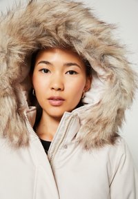 Parka beige clair avec une capuche en fausse fourrure dans des tons de brun clair. Dispose d'une fermeture éclair et de pressions ; texture de tissu lisse.