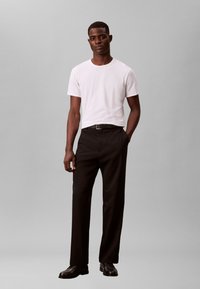 Λευκό t-shirt με στρογγυλή λαιμόκοψη και στενό μαύρο παντελόνι με ζώνη. Το μοντέλο στέκεται άνετα, φορώντας μαύρα δερμάτινα παπούτσια. Γκρι φόντο.