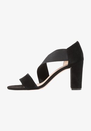 Sandal de tacón alto de ante negro con una amplia correa elástica a través del arco y una plantilla acolchada. Presenta un tacón en bloque grueso.