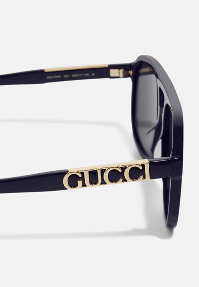 Lunettes de soleil en plastique noir avec un audacieux logo doré "GUCCI" sur la branche, d'une forme rectangulaire élégante et de verres teintés.
