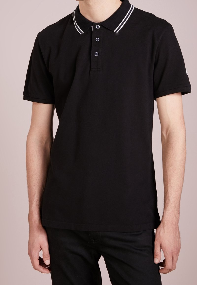 T-shirt polo noir en coton, avec un col classique orné de deux rayures blanches, une patte de boutonnage à trois boutons et des manches courtes.