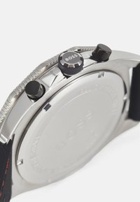 Reloj de acero inoxidable con acentos negros, que presenta dos botones de presión y una corona texturizada. Parte trasera plateada con un acabado suave.