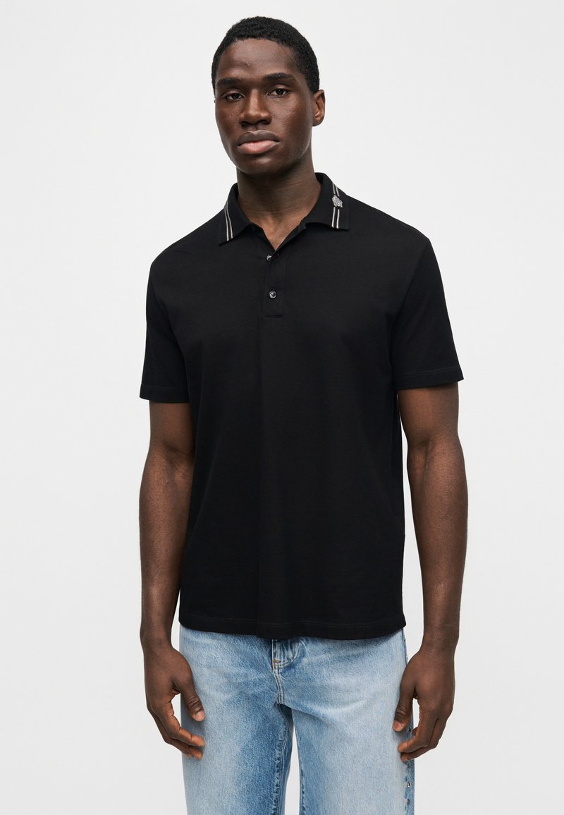 Homme portant un polo noir à manches courtes avec un col à rayures, et un jean bleu clair, debout devant un fond blanc uni.