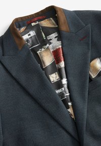 Blå struktureret blazer med brune detaljer ved kraven og lommerne, med et mønstret indre, der viser vintage kameraer og flasker.