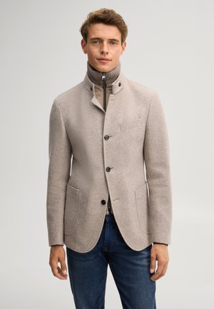 Jonge man die een beige gestructureerde blazer draagt over een lichtbruine coltrui met rits en een blauwe spijkerbroek, staand tegen een effen achtergrond.