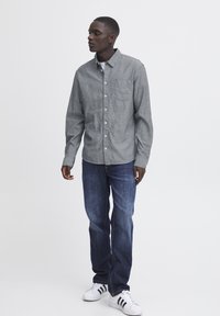 Camisa de botones a rayas grises con un solo bolsillo en el pecho, combinada con pantalones vaqueros oscuros y zapatillas blancas con rayas negras.