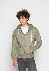Grijze hoodie met ritssluiting van een zachte, gestructureerde stof met een capuchon, trekkoorden en vo pockets. Gepaard met een wit shirt en grijze jeans.