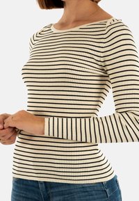 Donna che indossa un maglione a coste crema con strisce orizzontali nere e jeans blu, ripresa dalla nuca ai fianchi, con le mani unite.