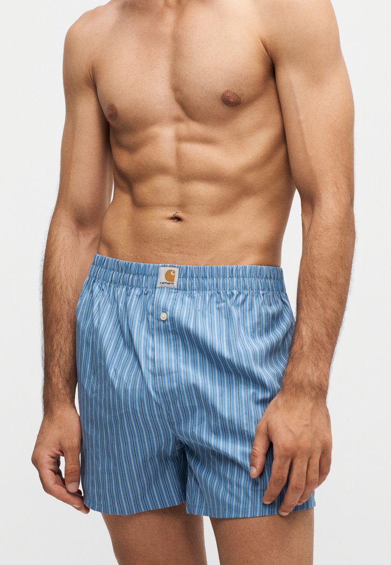 Hemdloser Mann trägt blaue und weiße vertikal gestreifte Carhartt Boxershorts mit elastischem Bund und vorderem Knopfverschluss vor einfarbigem Hintergrund.