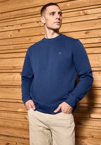 Marineblå sweatshirt i bomullsblandning, rundad halsringning, ribbade ärmslut och nederkant, i kombination med ljusbeiga byxor, mot en träbakgrund.