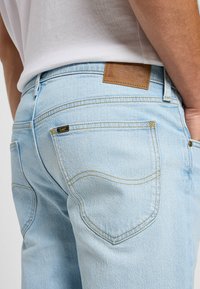 Jeans de mezclilla azul claro con un acabado texturizado, que presentan costuras amarillas y un parche de cuero marrón con logotipo en relieve en la cintura.