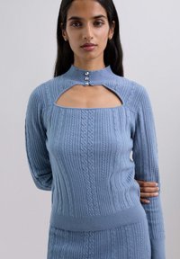 Suéter de punto azul claro con un cuello alto, textura acanalada y un diseño con corte en el pecho. Presenta tres botones decorativos.