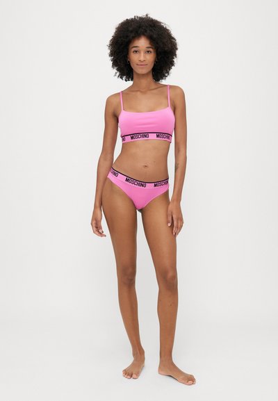 Un bralette rose et un slip assorti arborent une bande logo Moschino noire. Le tissu semble lisse avec un design ajusté et des fines bretelles.
