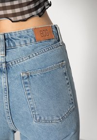 Vaqueros de mezclilla azul con un diseño de tiro medio, una etiqueta de cuero marrón marcada "BDG" y un bolsillo trasero visible con detalles de costura.