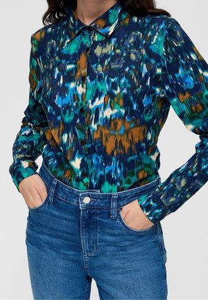 Woman wearing buttoned long-sleeve shirt with blue, green, white, and brown abstract pattern tucked into blue denim jeans with hands in pockets.