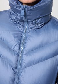 Gros plan sur une personne portant une veste matelassée bleu clair brillant avec un col haut et une fermeture éclair centrale partiellement zippée.