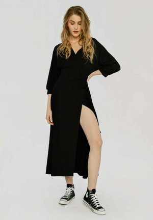 Donna con lunghi capelli biondi che indossa un vestito nero avvolgente con uno spacco alto e sneakers nere alte, in piedi davanti a uno sfondo semplice.