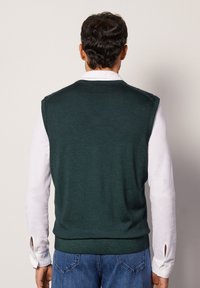 Zielony bezrękawnik o gładkiej fakturze, z ribbroadź i luźnym kroju, nałożony na białą koszulę z długim rękawem. Swobodne dżinsy.