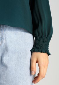 Blusa verde testurizzata con maniche arricciate, caratterizzata da un polsino a volant. Abbinata a jeans in denim azzurro, che mostrano una vestibilità comoda.