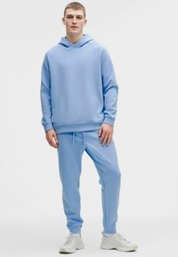 Světle modrá mikina s kapucí, klokáním kapsou, žebrovanými manžetami a lemem, doplněná o odpovídající tepláky. Látka působí měkce a pohodlně.