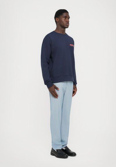 Felpa navy con logo a patch sul davanti, abbinata a pantaloni a righe blu chiaro e scarpe nere slip-on. Tessuto morbido, vestibilità rilassata.