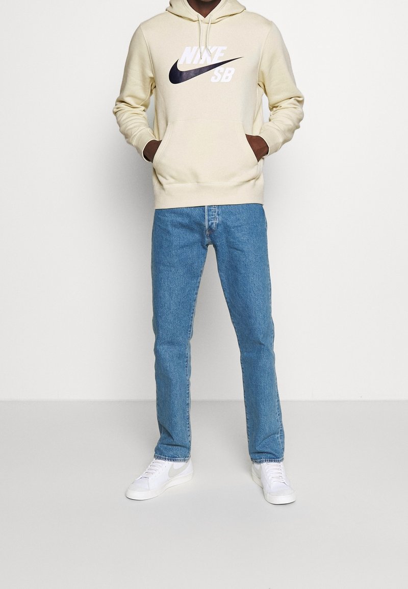 Person i krämfärgad Nike SB-hoodie, blå jeans och vita Nike-sneakers, stående med händerna i hoodiefickorna mot en enfärgad bakgrund.