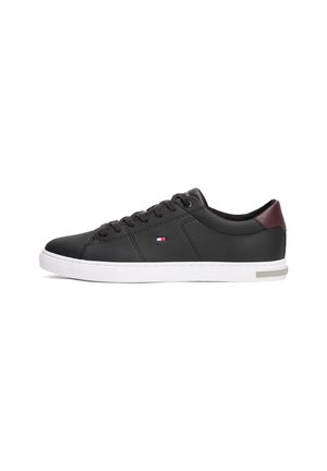 Schwarzer Low-Top-Sneaker mit weißer Sohle, schwarzen Schnürsenkeln, kleinem Flaggenlogo an der Seite und dunkelrotem Fersenpatch.