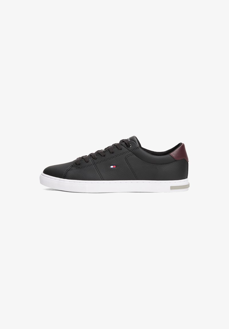 Baskets basses noires avec semelle blanche, lacets noirs, petit logo drapeau sur le côté et patch rouge foncé au talon.