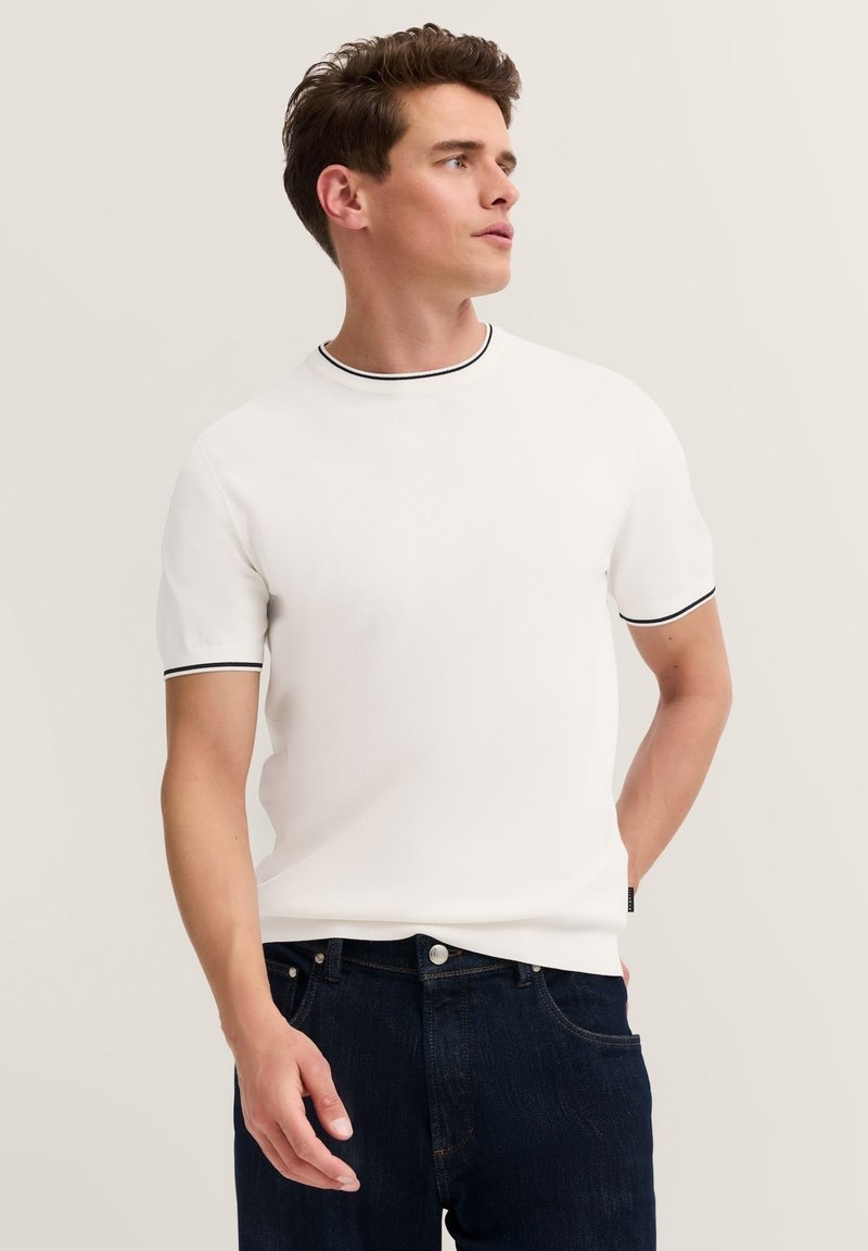 Weißes T-Shirt mit kurzen Ärmeln und schwarzem Besatz am Kragen und an den Ärmeln, gefertigt aus einem strukturierten Stoff, kombiniert mit dunkelblauen Jeans.