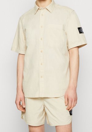 Homme portant une chemise beige à manches courtes avec poche poitrine et un short assorti, tous deux ornés de patches logo Calvin Klein noirs.