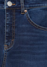 Jeans de denim azul oscuro con costuras doradas en contraste, que presentan un botón de metal y detalles en los bolsillos laterales. Textura suave y diseño clásico.
