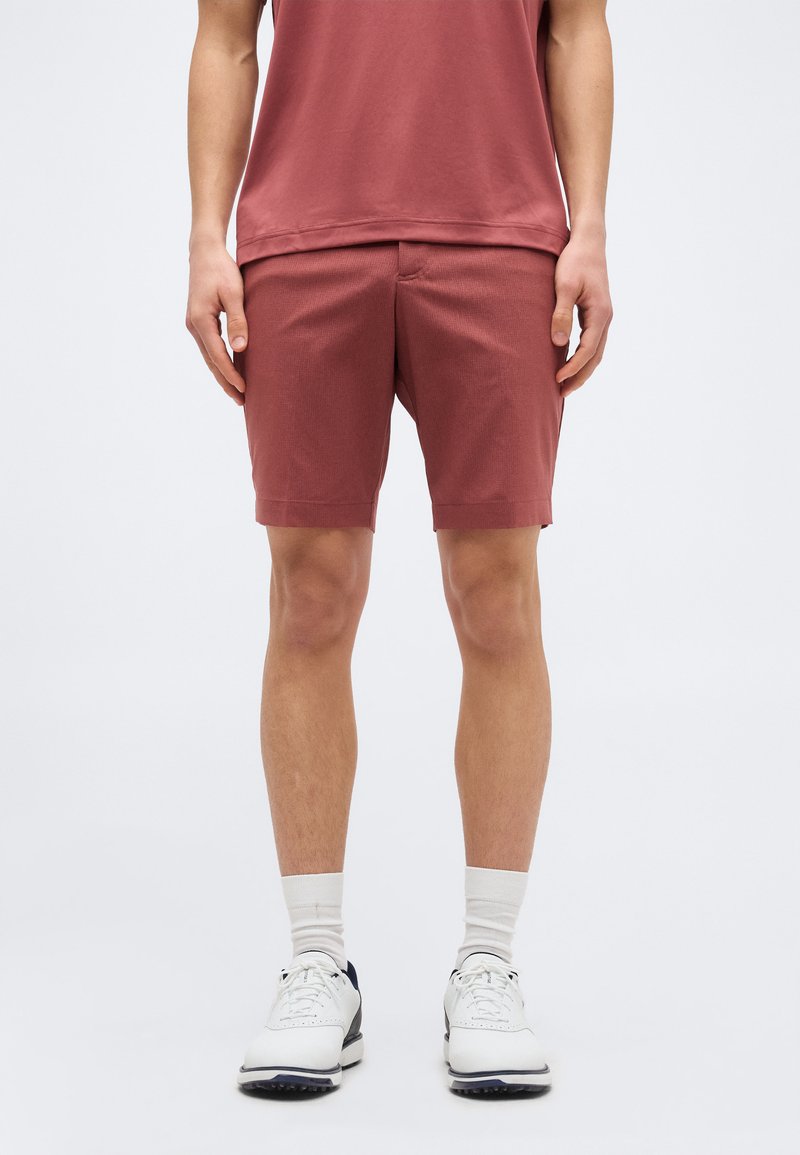 Mann trägt rostfarbene Shorts, passendes Oberteil, weiße Socken und weiße Sportschuhe und steht vor einem einfarbigen Hintergrund.