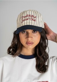 Creme baseball cap med røde lodrette striber og "Prohibited Social Club" broderet i rød, med en marineblå skygge og justerbar bagside.