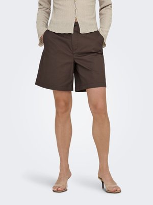 Person i mørkebrune shorts til kneet, beige teksturert knappet topp og gjennomsiktige sandaler med hæl, står med hendene i lommene.